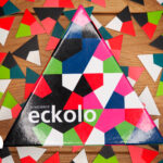 Das Anlegespiel hat eine dreieckige Spielbox mit dreieckigen bunten Elementen. Im unteren Drittel steht der Name eckolo in weißen Kleinbuchstaben. Im Hintergrund liegen durcheinander gewürfelte bunte Dreiecke auf einem mittelbraunen Holztisch.