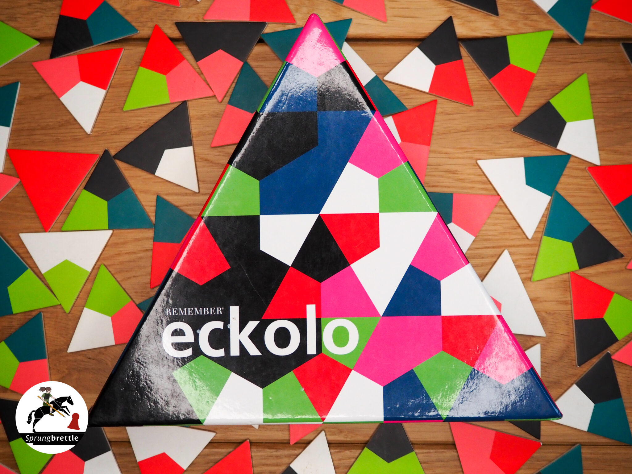 Das Anlegespiel hat eine dreieckige Spielbox mit dreieckigen bunten Elementen. Im unteren Drittel steht der Name eckolo in weißen Kleinbuchstaben. Im Hintergrund liegen durcheinander gewürfelte bunte Dreiecke auf einem mittelbraunen Holztisch.