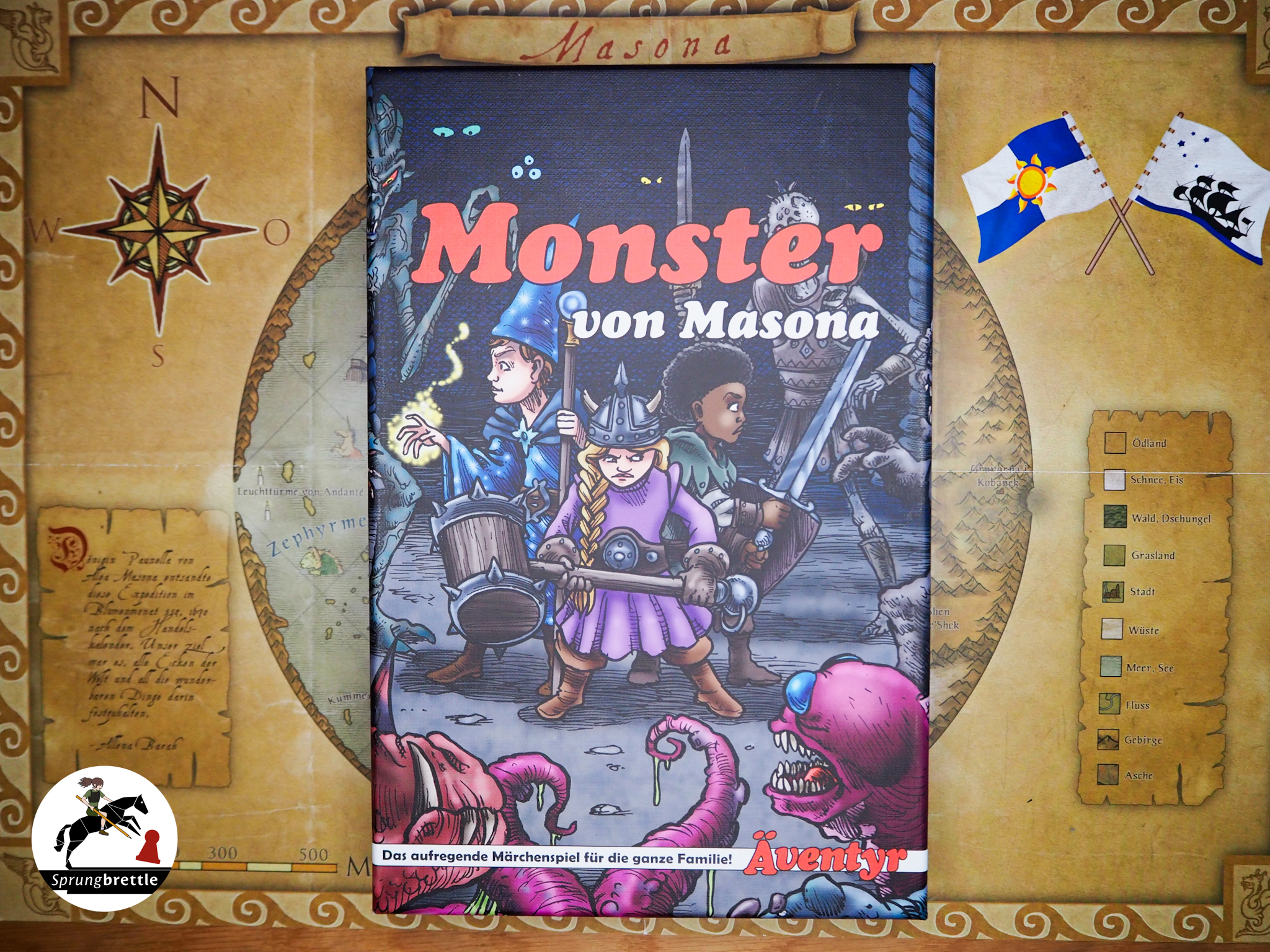 Die Spielbox der Erweiterung Monster von Masona liegt mittig auf einer bräunlichen Landkarte. Farblich ist sie dunkel gehalten mit schwarzem Hintergrund. Im unteren Drittel ist ein dunkel lila Monste mit Tentakeln, großen Zähnen und blauen Glubschaugen zu sehen. Mittig im Bild stehen drei Abenteurerinnen und Abenteurer mit gezückten Waffen Rücken an Rücken um die sie umkreisenden Untoten abzuwehren.