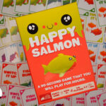 Partyspiel Happy Salmon schräg in der Mitte. Hauptsächlih orange mit einem neongelben Fisch in der Mitte. Im Hintergrund ein Haufen Spielkarten.
