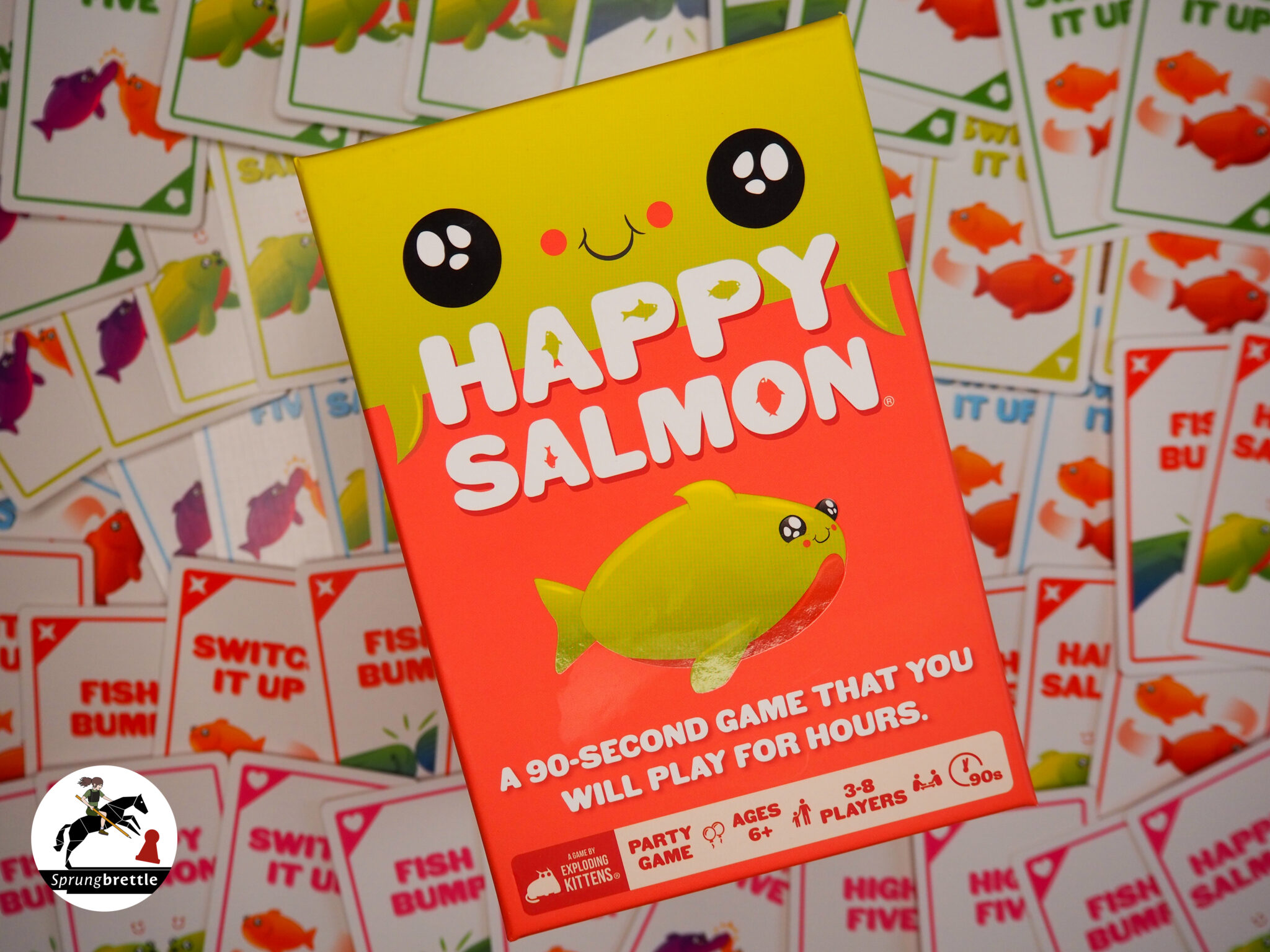 Partyspiel Happy Salmon schräg in der Mitte. Hauptsächlih orange mit einem neongelben Fisch in der Mitte. Im Hintergrund ein Haufen Spielkarten.