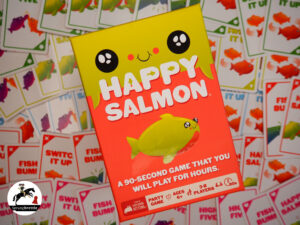 Partyspiel Happy Salmon schräg in der Mitte. Hauptsächlih orange mit einem neongelben Fisch in der Mitte. Im Hintergrund ein Haufen Spielkarten.