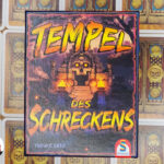 Deduktionsspiel Tempel des Schreckens mittig. Orange Gelber Titel in Großbuchstaben. Im Hintergrund ein Tempeleingang mit einer Totenkopftür mit leuchtend gelben Augen.